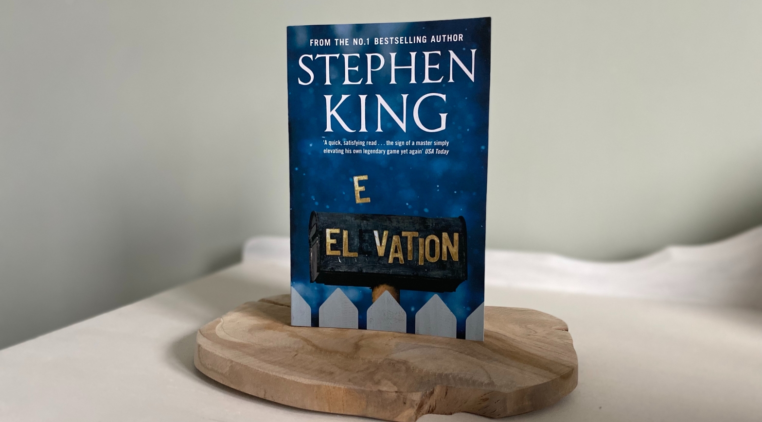 The book Elevation by Stephen King standing on a wooden platform, in front of a green wall.